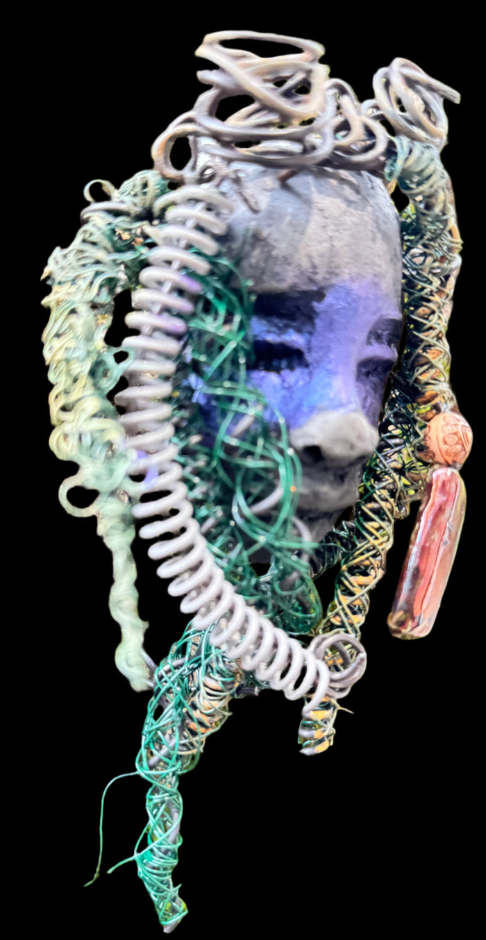 Rayda is a 4” x 6” 5oz. mixed media raku fire mask that includes 15 feet of 24 and 16 gauge wire. It has a Smokey black two tone complexion and a large metallic Raku fired earring with a wooden bead. Its earthy appeal is enhanced when displayed with natural tree bark, making it a lovely addition to your HerDew mask collection.