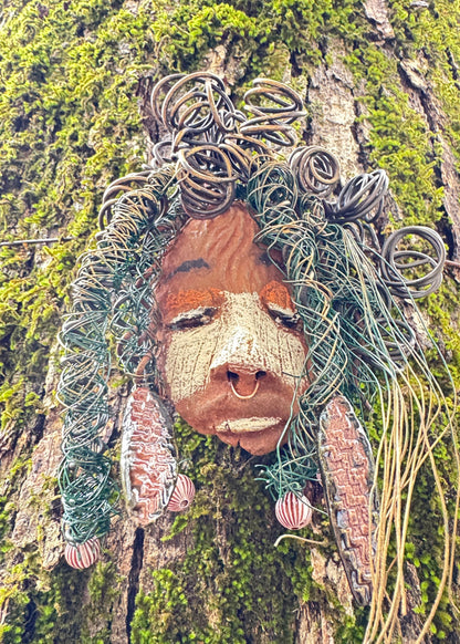 Baldwin Bo is a mixed media raku fire mask, measuring 4" x 6" and weighing 6oz. This unique piece features 25 feet of 24 and 16 gauge wire and a copper nose ring, giving it a three-colored complexion. The mask is also adorned with large Raku fired earrings and a wooden bead, and looks stunning when photographed against a mossy tree bark. Add Baldwin Bo to your collection of HerDew masks for a beautiful and eye-catching addition.