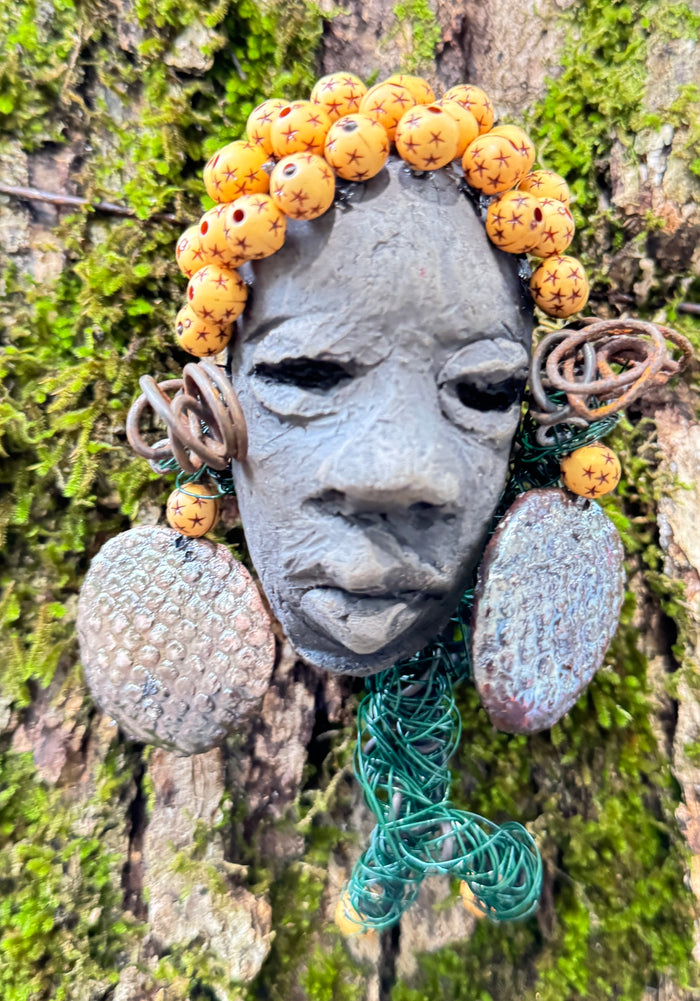 Introducing Cali, a 3” x 5” 5oz. mixed media raku fire mask adorned with a copper nose ring, three-colored complexion, and two large Raku fired earrings with a wooden bead. With 15 feet of 24 and 16 gauge wire, this piece is a stunning addition to your HerDew mask collection. See the beautiful Cali against a tree bark background.