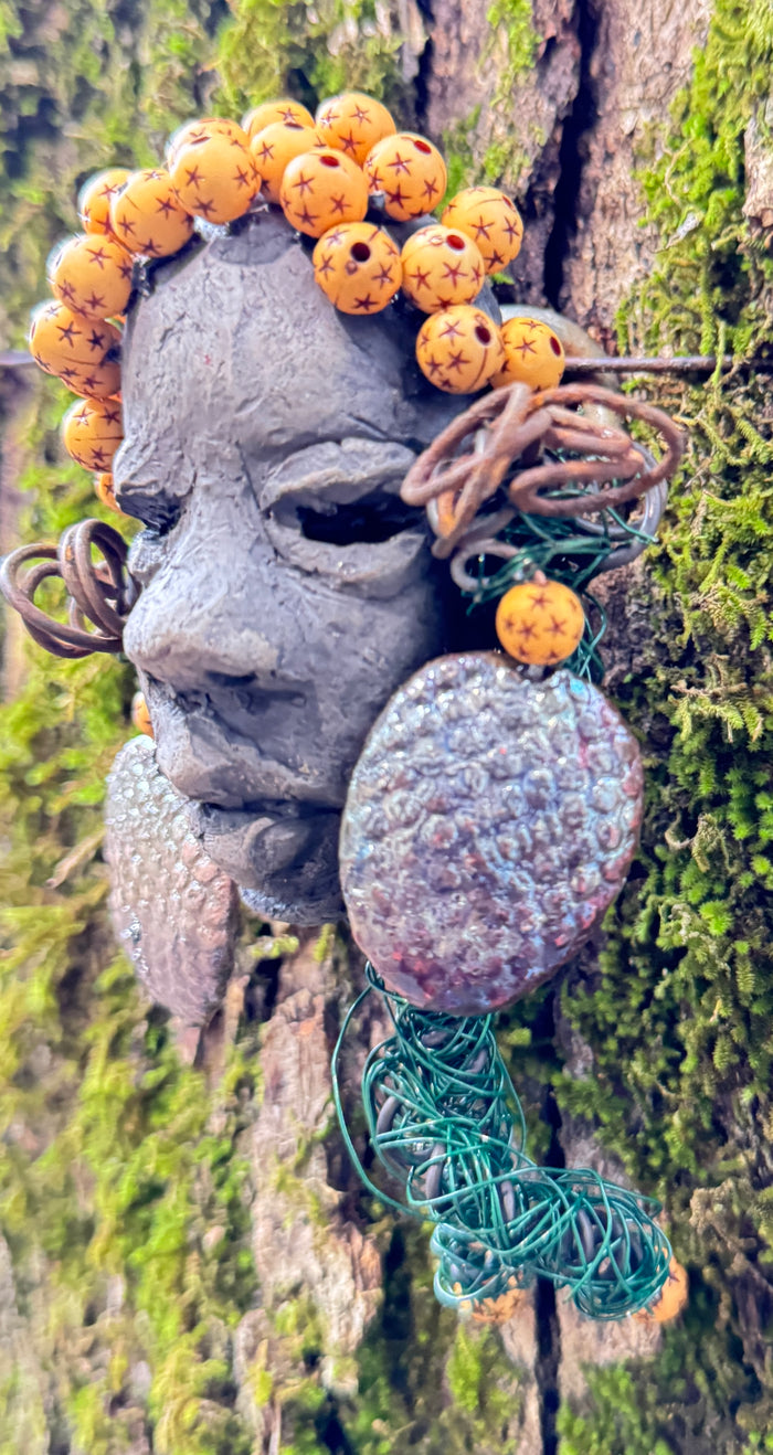 Introducing Cali, a 3” x 5” 5oz. mixed media raku fire mask adorned with a copper nose ring, three-colored complexion, and two large Raku fired earrings with a wooden bead. With 15 feet of 24 and 16 gauge wire, this piece is a stunning addition to your HerDew mask collection. See the beautiful Cali against a tree bark background.
