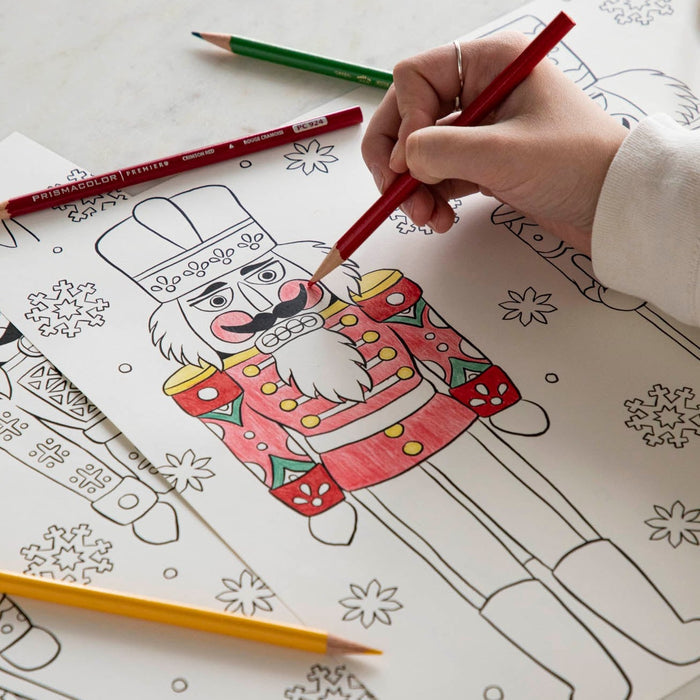 A woman coloring a nutcracker on the Christmas Coloring Placemat with scattered colored pencils.
