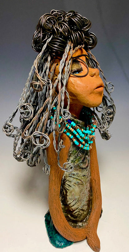 Meet Clarice! Clarice stand 13" x 6" x 5" and weighs 4.03 lbs. She has a lovely honey brown complexion with big bright eyes. Clarice wears glasses now! Her Herdew took over 2 hours to complete. It's made with 16 gauge wire. Clarice's dress is alligator green with streaks of copper. She has a blue beaded spiral necklace around her neck. Clarice looks to have a New ExtrArtindary Vision for 2021!