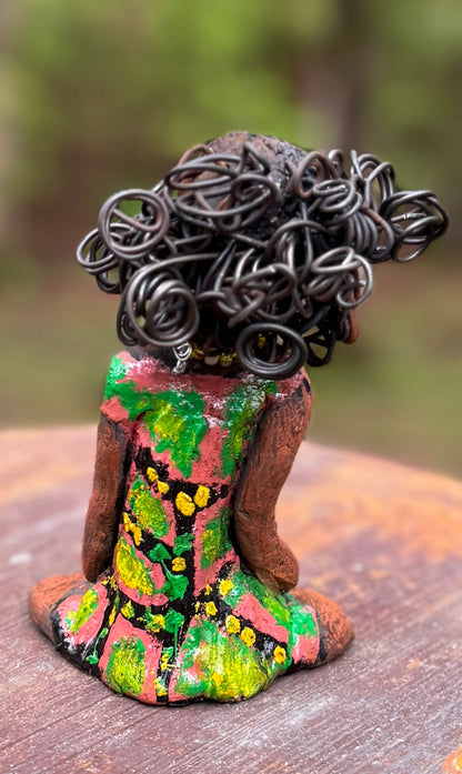 Introducing Katie, the fearless figurine who measures 6" x 4" x 3" and weighs a mere 11 ozs. Donning a stunning speckled red and green dress and with her coiled hair as her crown of glory, Katie exudes confidence and courage as she sits in a yoga pose, exuding curiosity. Begin your journey with the Herdew Collection and experience the thrill of free shipping and our customer-friendly return policy. Don't miss out on this one-of-a-kind piece!