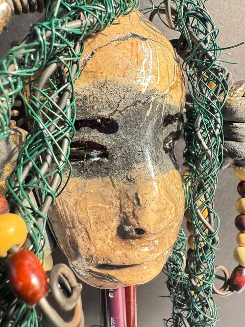 Decorative mixed media raku fired  mask  surrounded with green hair and wooden beads on a gray background